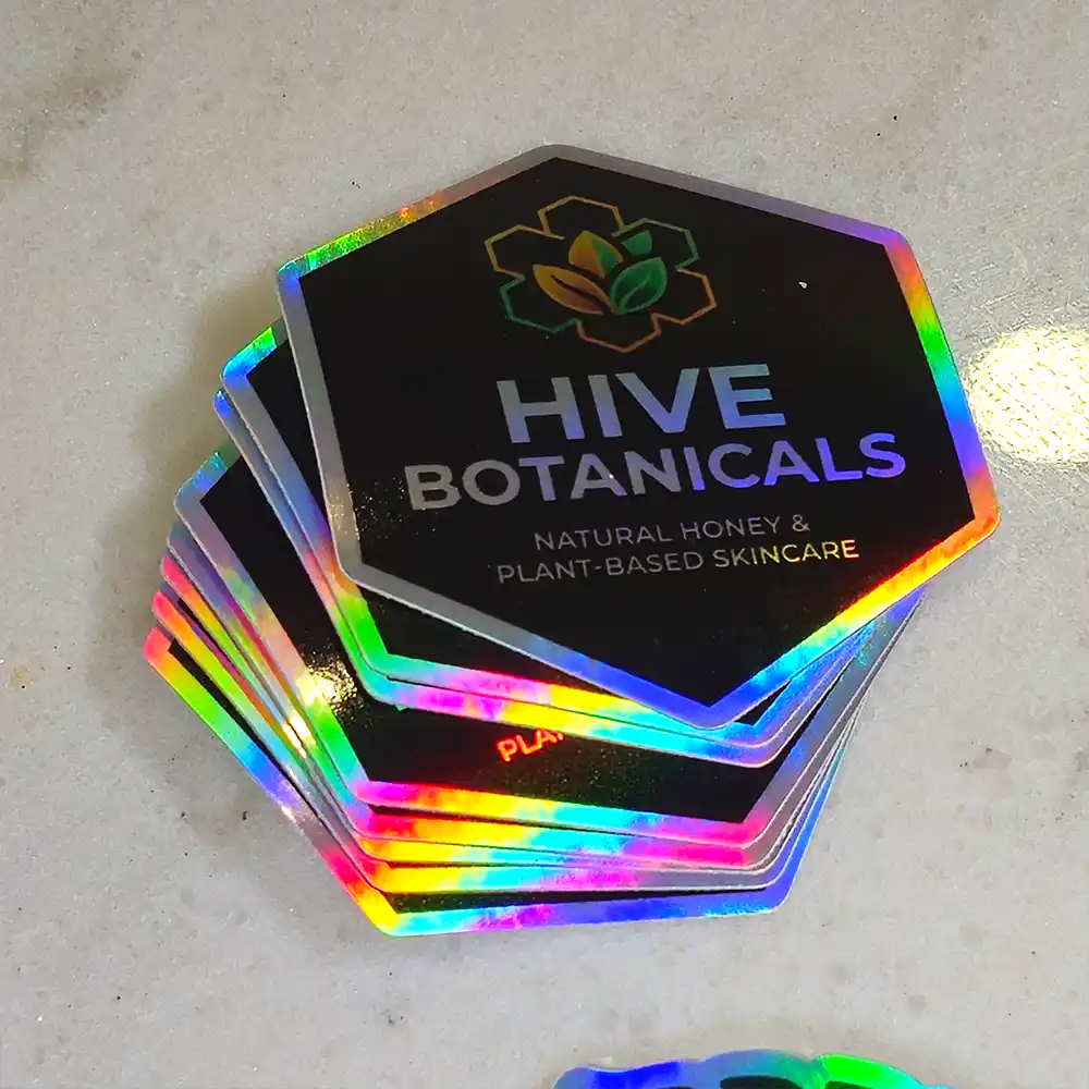 holographic-promo-stickers-2 • StickerPlanet Canada Stack of black holographic hexagonal tags with Hive Botanicals logo and text: Natural Honey & Plant-Based Skincare (glittering rainbow edges).