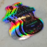 holographic-promo-stickers-4 • StickerPlanet Canada Holographic guitar-shaped stickers stacked, displaying 'Timber & Tone Instruments' with colorful rainbow edges and floral design