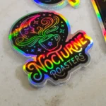 holographic-promo-stickers-5 • StickerPlanet Canada Holographic sticker with a steaming coffee cup and 'Nocturne Roasters' in rainbow lettering.