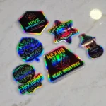 holographic-promo-stickers-6 • StickerPlanet Canada Six iridescent patches with company logos on a light surface, including Hive Botanicals, Nova Kinetic Robotics, Flora Moss Plant Studio, Nexus Heavy Industries, Nocturne Roasters, and Timberstone Instruments.