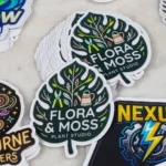promo-stickers-2 • StickerPlanet Canada Round leaf-shaped sticker for Flora & Moss Plant Studio with botanical icons (watering can, pruning shears) and white text on dark green background.