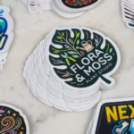 promo-stickers-3 • StickerPlanet Canada Stack of round stickers featuring a botanical design and the text 'Flora & Moss Plant Studio' on a dark green background in the center group of stickers.