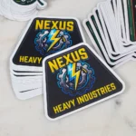 promo-stickers-4 • StickerPlanet Canada Sticker with Nexus Heavy Industries logo: black octagonal badge, yellow text, a gear and blue lightning bolt in the center.