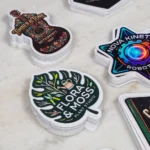 promo-stickers-7 • StickerPlanet Canada Assorted die-cut stickers on a light surface, including a leafy 'Flora Moss Plant Studio' design, a guitar-themed 'Timber & Tone Instruments' sticker, and a colorful 'Nova Kinet' robot graphic.